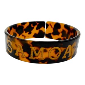Puede incluir: Un brazalete ancho y abierto con un patrón de carey en tonos marrones y negros. La palabra SAMOA está grabada en letras mayúsculas de color dorado en la parte frontal del brazalete, que tiene un acabado liso y pulido y una forma curva.