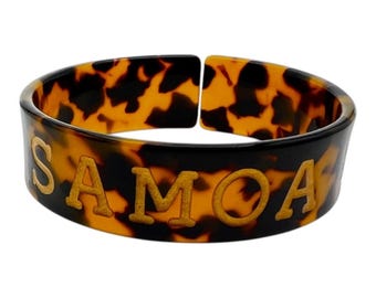 Puede incluir: Un brazalete ancho y abierto con un patrón de carey en tonos marrones y negros. La palabra SAMOA está grabada en letras mayúsculas de color dorado en la parte frontal del brazalete, que tiene un acabado liso y pulido y una forma curva.