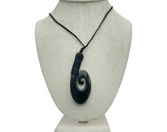 Puede incluir: Un collar con colgante tallado en verde oscuro en forma de espiral, expuesto en un maniquí. El colgante está suspendido de un cordón negro. El cordón está unido al colgante con una sección envuelta. El collar es una joya única.