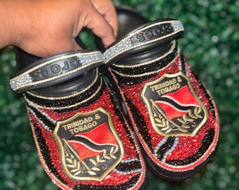 Könnte beinhalten: Ein Paar schwarze Crocs, verziert mit roten, schwarzen und goldenen Strasssteinen. Die Crocs haben ein goldenes Schild mit dem Text "Trinidad & Tobago" und der Flagge von Trinidad und Tobago.