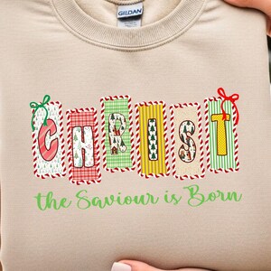 May include: A beige sweatshirt with the word "CHRIST" in colorful, patterned blocks. The text "the Saviour is Born" is below in green. The sweatshirt is held up by a person.