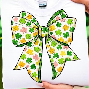 May include: White sweatshirt featuring a large bow design with green, yellow, and pink shamrocks. The bow has a green outline and is centered on the front of the shirt. The shirt is held up by a person.