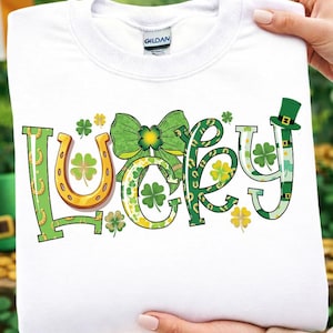 Puede incluir: Sudadera blanca con la palabra "Lucky" en letras verdes, amarillas y doradas. La "L" y la "Y" están decoradas con herraduras y tréboles. Un lazo verde y un sombrero de duende completan el diseño del Día de San Patricio.