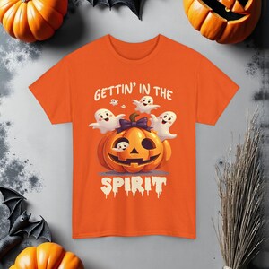 Puede incluir: Camiseta naranja con un diseño de Halloween. El gráfico muestra una calabaza sonriente, fantasmas y el texto "GETTIN' IN THE SPIRIT". La imagen está decorada con calabazas y murciélagos, creando un tema festivo de Halloween.