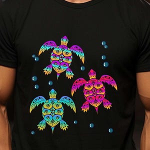May include: Black t-shirt featuring three colorful sea turtle designs. Each turtle has a unique pattern with shades of blue, yellow, pink, and purple. Blue bubbles are scattered around the turtles.
