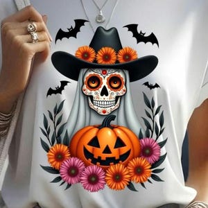 以下が含まれることがあります： ハロウィーンをテーマにしたデザインの白いTシャツ。グラフィックには、オレンジ色の花で飾られた黒い帽子をかぶったシュガースカル、彫刻されたカボチャ、コウモリが含まれています。デザインはオレンジとピンクの花に囲まれています。