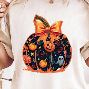 May include: Cream-coloured t-shirt with a large pumpkin design. The pumpkin is black with orange details, including a bow, smaller pumpkins, stars, and a ghost. The design has a Halloween theme.