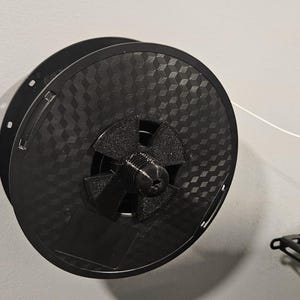 以下が含まれることがあります： 白い壁に取り付けられた黒い3Dプリンターフィラメントスプールホルダー。 スプールホルダーは、テクスチャード加工された幾何学模様と、黒いデバイスに向かって伸びる白いフィラメントストランドを備えています。