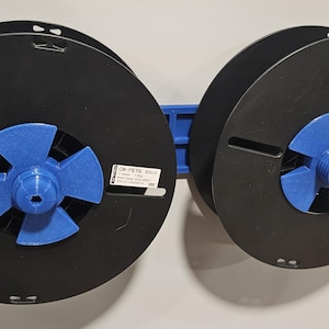 May include: Two black spools with blue centers are connected by a blue bracket. The spools have a label on the side. The spools are mounted on a white wall. The spools are likely for ribbon or tape.