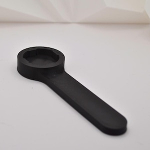 Puede incluir: Una herramienta negra y gomosa con una cabeza circular y un mango largo y plano. La cabeza tiene un interior empotrado y multifacético. La herramienta está diseñada para un propósito específico, probablemente relacionado con apretar o aflojar algo.