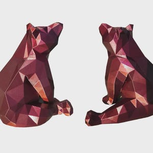May include: Two geometric bear sculptures in shades of burgundy and pink sit side-by-side. The bears are in a seated position, with their legs extended. The sculptures have a faceted, polygonal design, giving them a modern look.