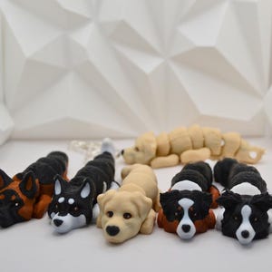 May include: A collection of dog figurines in various breeds, including a German Shepherd, Husky, and Golden Retriever. The figurines are in shades of black, brown, and tan, with detailed features. The dogs are arranged on a white surface.
