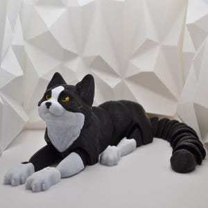 Puede incluir: Una figura de gato blanco y negro con ojos amarillos, tumbada con la cola enroscada. El gato tiene patas blancas y pecho blanco. La figura está sobre un fondo geométrico blanco.