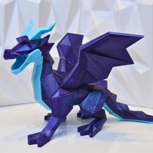 以下が含まれることがあります： 青い頭と翼、紫色の体を持つ3Dプリントされたドラゴンフィギュア。ドラゴンは、質感のあるキラキラした仕上げになっています。