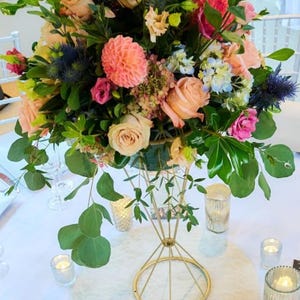 May include: A vibrant floral centerpiece with pink, peach, and white roses, blue thistle, and greenery. The arrangement sits in a gold geometric vase on a white tablecloth, with candles and glassware. A table number sign is also present.