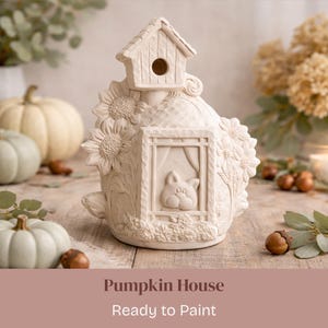 May include: A cream-colored pumpkin-shaped house with a birdhouse on top, sunflowers, and a cat in a window. The house is ready to paint and is surrounded by pumpkins and acorns.