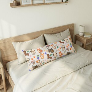 May include: A long rectangular body pillow featuring a repeating pattern of various cartoon dog breeds, colorful paw prints, bones, and pet bowls on a white background. The pillow is positioned horizontally across a bed with neutral bedding.