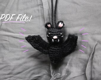 Op de afbeelding: Een zwarte gehaakte vleermuis met roze oren en tanden die aan een zwart koord hangt. De vleermuis bevindt zich op een grijze stoffen achtergrond met de tekst "PDF File!" in wit.