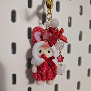 May include: A small flocked animal figurine wearing a red and white polka dot outfit with a white hood and red ribbon. The figure is attached to a gold-tone lobster clasp keychain featuring a dangling charm strand with red bows, clear beads, and stars.