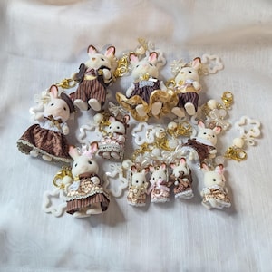 May include: A collection of miniature rabbit figurines, each dressed in detailed outfits. The rabbits are in various poses, some with accessories, and attached to keychains with decorative charms. The color palette includes browns, golds, and whites.