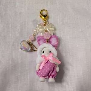 May include: A keychain featuring a small white animal figure wearing a pink polka-dot hooded outfit with a pink ribbon. The gold-tone clasp is adorned with a white bow, a pink flower bead, a small heart-shaped charm, and gold angel wings.