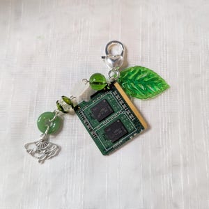 Puede incluir: Un llavero único con una placa de circuito verde, adornado con cuentas verdes, un dije de abanico plateado y un cierre en forma de corazón. Una hoja verde y una cuenta blanca se suman al diseño. Un accesorio creativo.