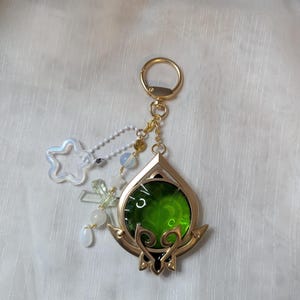 May include: A gold-coloured keyring with a green, teardrop-shaped charm. The charm has a gold frame and a green centre. Attached are a star-shaped charm, beads, and a gold keyring.