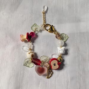 May include: A whimsical charm bracelet featuring a gold chain and clasp. The bracelet is adorned with various charms, including a red apple, a heart, a white bunny, and leaf-shaped beads. The charms are in shades of red, white, and gold.
