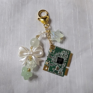 Puede incluir: Un llavero dorado con un cierre en forma de corazón, con un lazo blanco, cuentas verdes y transparentes, una flor verde y un pequeño dije de placa de circuito. El llavero también incluye un dije en forma de estrella.