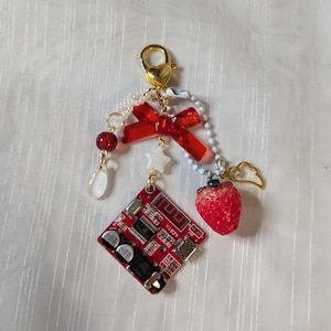 Puede incluir: Un llavero único con un dije de placa de circuito rojo, una fresa roja, un lazo rojo y un cierre dorado en forma de corazón. El llavero también incluye cuentas blancas, una estrella y un dije de ala dorada.
