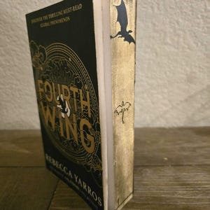 May include: A black hardback book with a gold spine and a dragon design. The book title is "Fourth Wing" and the author is "Rebecca Yarros".