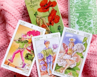 May include: A tarot card deck titled "The Spirit of Flowers Tarot" with a green box featuring a poppy illustration. Three tarot cards are visible, each with colorful illustrations of figures and flowers. The cards are on a pink crocheted blanket.
