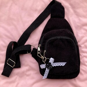 May include: A black corduroy crossbody bag with a black bow and white lace trim. The bag has a zipper closure and a small pocket on the front.