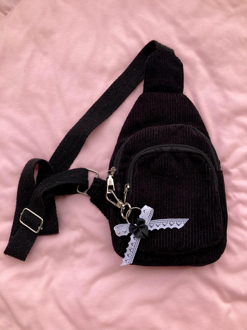 May include: A black corduroy crossbody bag with a black bow and white lace trim. The bag has a zipper closure and a small pocket on the front.