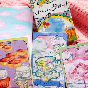 Może przedstawiać: Kolorowa talia kart tarota ze słowami "Day Dream Tarot" na pudełku. Widocznych jest kilka kart tarota, z fantazyjnymi ilustracjami szefa kuchni, filiżanki do herbaty i flaminga. Karty są umieszczone na różowym, teksturowanym tle.
