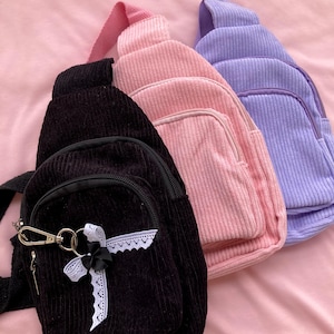 May include: Three corduroy crossbody bags in black, pink, and lavender. The black bag has a white lace trim and a silver charm.