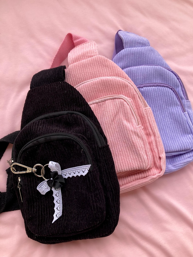 May include: Three corduroy crossbody bags in black, pink, and lavender. The black bag has a white lace trim and a silver charm.