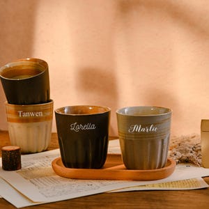 May include: Ceramic cups in brown, beige, and olive green. Some are stacked, others on a wooden tray. The names "Tanwen", "Lorella", and "Marlie" are inscribed on the cups. The scene is set on a table with a vintage postcard.