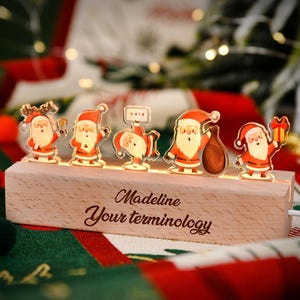 Może przedstawiać: Drewniana ekspozycja świetlna bożonarodzeniowa z pięcioma figurkami Świętego Mikołaja. Podstawa jest grawerowana napisem "Madeline Your terminology". Figurki są podświetlane, tworząc świąteczną scenę. Dekoracje świąteczne w tle.