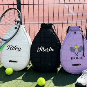 May include: Three tennis racket bags in white, black, and lavender, each with a quilted pattern and a name printed on the front. A tennis racket and two tennis balls are next to the bags. A scoreboard shows the number zero.