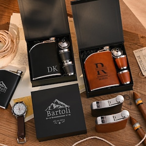 May include: A selection of flasks, shot glasses, and a watch, displayed in black gift boxes. One flask is black with initials, another is brown with a name and date. The watch has a brown leather strap. Cigars and a tag are also present.