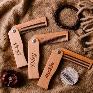 Könnte beinhalten: Eine Sammlung personalisierter, zusammenklappbarer Holzkämme, jeweils mit einem Namen graviert: "Bride", "Riley" und "Jackie". Ein dunkelbraunes Lederarmband und ein kleiner, runder Behälter sind ebenfalls vorhanden. Die Artikel liegen auf einer strukturierten, beigefarbenen Oberfläche.