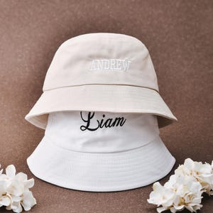 Könnte beinhalten: Zwei weiße Bucket-Hats mit schwarzer Stickerei. Der obere Hut sagt "ANDREW" und der untere Hut sagt "Liam".