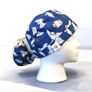May include: A blue scrub cap featuring a pattern of white owls carrying letters. The cap has a gathered back and a cream-colored tie. The owls are depicted in flight, with some holding envelopes.