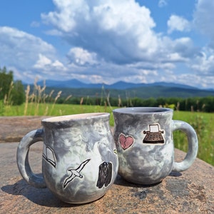 May include: Two gray ceramic mugs with a marbled glaze. One mug has a book, a bird, and a suit drawn on it. The other mug has a heart, a typewriter, and a bird drawn on it.