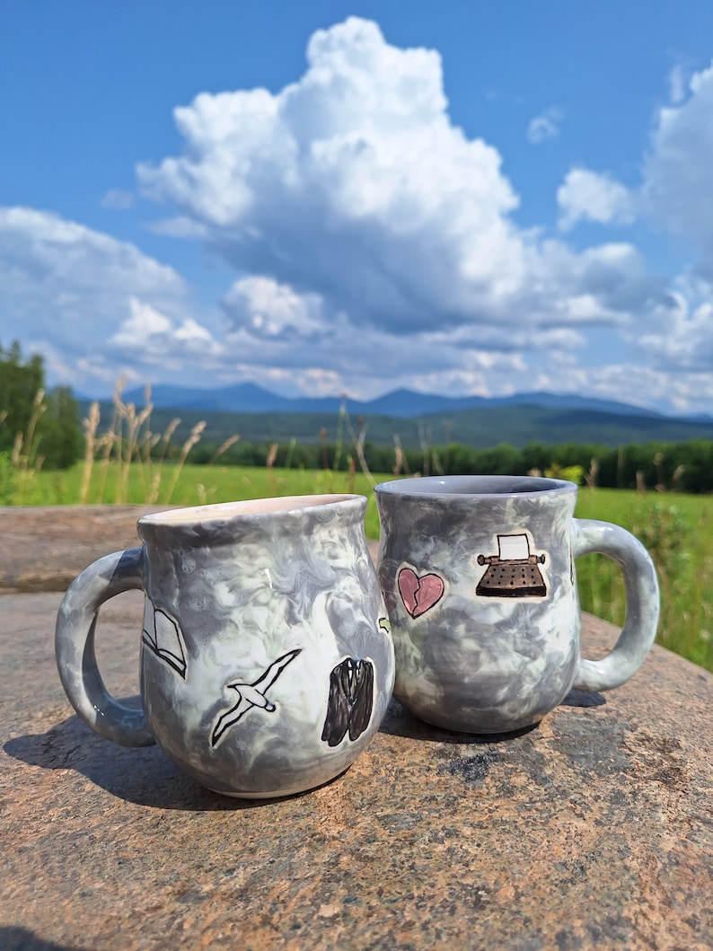 May include: Two gray ceramic mugs with a marbled glaze. One mug has a book, a bird, and a suit drawn on it. The other mug has a heart, a typewriter, and a bird drawn on it.