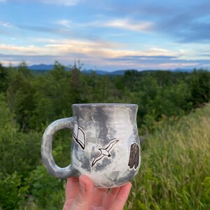 May include: A gray ceramic mug with a white glaze and a marbled effect. The mug has a handle and is decorated with a black and white illustration of a book, a bird, and a suit.