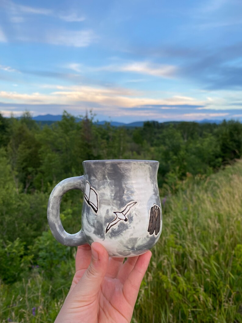 May include: A gray ceramic mug with a white glaze and a marbled effect. The mug has a handle and is decorated with a black and white illustration of a book, a bird, and a suit.