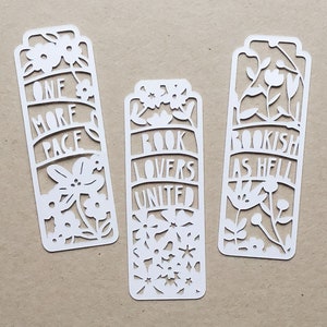 May include: Three white paper bookmarks with floral designs and text. The bookmarks read "One More Page", "Book Lovers United", and "Bookish As Hell".