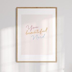 以下が含まれることがあります： 「You beautiful Nerd」の文字が筆記体で書かれた額入りのアートプリント。文字はピンク、オレンジ、ブルーで、薄いピンク色の背景に配置されています。フレームは薄茶色です。